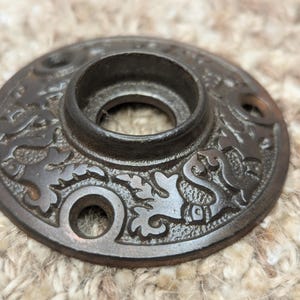 May include: A round, black metal door knob with an ornate design featuring a floral pattern and a central hole. The knob has a slightly raised edge and a smooth, polished finish.