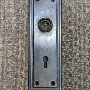 May include: A silver door plate with a keyhole and a round knob. The plate has a rectangular shape with rounded corners and is decorated with four small decorative circles.