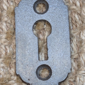 Könnte beinhalten: Ein dunkelgrauer Schlüsselschlitzabdeckung aus Metall mit zwei runden Löchern und einem Schlüsselschlitz in der Mitte.