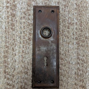 Op de afbeelding: Een rechthoekige, verweerde bruine metalen deurplaat met een centrale ronde opening voor een deurknop en een sleutelgat aan de onderzijde. De plaat heeft afgeschuinde randen en vier kleine schroefgaten, afgewerkt met een donkere, geoxideerde laag.