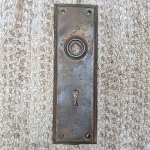 Pode incluir: Uma placa de porta retangular de latão desgastado com uma abertura circular central para o eixo e uma fechadura abaixo. A superfície metálica apresenta uma pátina escura e envelhecida com sinais visíveis de oxidação e desgaste, incluindo quatro orifícios para parafusos.