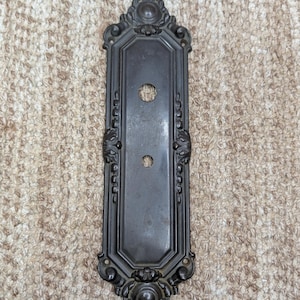 May include: A dark bronze-toned metal door plate featuring an elongated octagonal center panel with two circular holes. The plate is framed by ornate, symmetrical scrollwork and decorative finials at the top and bottom, with four small screw holes.