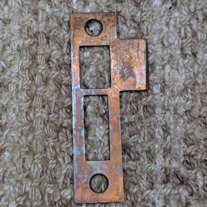 Peut inclure: Une gâche de porte rectangulaire en métal avec une finition cuivrée patinée. La plaque présente deux trous de vis circulaires en haut et en bas, une découpe carrée et une découpe rectangulaire, avec une languette saillante sur le côté droit.
