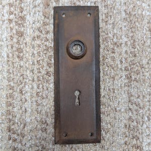 Pode incluir: Uma placa de porta retangular de bronze desgastado com uma abertura circular central para um puxador e uma fechadura abaixo. A superfície metálica apresenta uma pátina escura e envelhecida com quatro pequenos orifícios para parafusos.