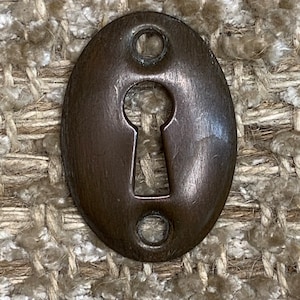Könnte beinhalten: Ein ovaler, antiker Schlüsselschlitzabdeckung aus dunkelbraunem Metall. Er hat zwei kleine Löcher auf beiden Seiten des Schlüsselloches.