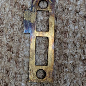 Peut inclure: Une gâche de porte rectangulaire en métal de couleur laiton avec une finition ternie. Elle comporte une longue ouverture rectangulaire, deux trous circulaires et une petite extension rectangulaire. Le métal présente des teintes dorées, bleues et violettes.
