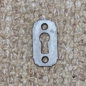 Op de afbeelding: Een kleine, rechthoekige, zilverkleurige sleutelgat afdekking met een decoratieve bovenrand. Het heeft een sleutelgatvorm in het midden, met een cirkelvormig gat erboven en eronder. Het metaal heeft een licht verouderd uiterlijk.