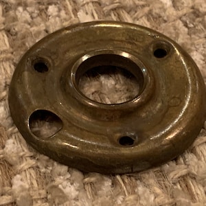 May include: A brass door knob backplate with four holes for screws. The backplate is round and has a slightly raised edge.
