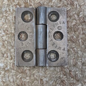 May include: A rectangular metal hinge with a central barrel and two flat plates, each featuring three countersunk screw holes. The metal surface is grey and weathered, with the word PHILADELPHIA embossed in a circular pattern around the holes.