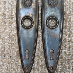 May include: Two antique brass door plates with a keyhole and a decorative design at the top. The plates are tarnished and have a worn look.