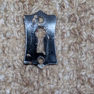 Pode incluir: Uma placa de metal preta para fechadura em forma de escudo, com uma abertura central para a chave e dois orifícios circulares para parafusos na parte superior e inferior. O acabamento apresenta sinais de desgaste com pequenas áreas de metal.