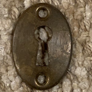 Op de afbeelding: Een vintage ovale sleutelgatbedekking van messing met twee kleine gaten aan beide zijden. De afdekking is licht aangetast en heeft een versleten uitstraling.