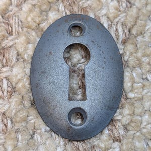 May include: A grey metal keyhole cover with two holes for mounting. The cover is oval shaped and has a keyhole cut out in the centre.