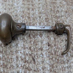 Op de afbeelding: Een vintage deurknopset van messing met een ronde knop aan het ene uiteinde en een gebogen hendel aan het andere uiteinde, verbonden door een vierkante metalen stift. Het beslag vertoont een verweerde patina met zichtbare schroeven.