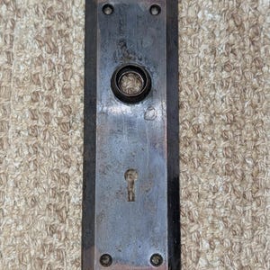 May include: A rectangular, dark-toned metal door plate featuring a beveled edge, a central circular opening for a knob, a lower keyhole cutout, and four small screw holes positioned at the top and bottom corners of the hardware piece.