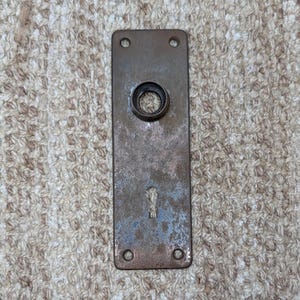 May include: A rectangular metal door plate with a weathered, brownish-grey patina. The plate features a central circular opening for a door knob spindle, a lower keyhole opening, and four small screw holes located at each of the four corners.