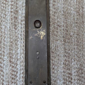 May include: A rectangular, weathered brass door plate featuring a central circular opening for a knob and a lower keyhole. The metal surface displays a dark, aged patina with visible signs of wear, scratches, and small screw holes at the top and bottom.