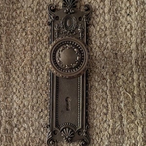 May include: Ornate antique bronze door hardware featuring a decorative oval doorknob and keyhole. The rectangular backplate has detailed embellishments, including a floral design at the bottom and a stylized top. The hardware is likely made of cast metal.