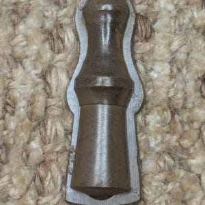 Op de afbeelding: Een bruine metalen flessenstop met een afgeronde bovenkant en een getextureerd oppervlak. De stop is in een metalen frame geplaatst.