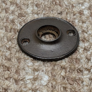 Pode incluir: Uma flange de piso circular de metal castanho escuro com uma abertura central roscada e dois pequenos orifícios de montagem nos lados opostos. A peça de ferragem tem um acabamento mate texturizado e uma secção central ligeiramente elevada.