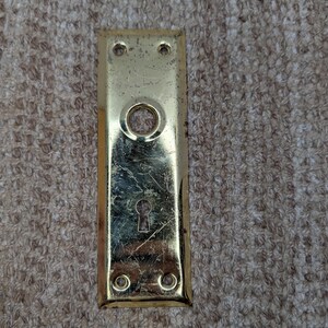 Puede incluir: Una placa de puerta rectangular de latón con acabado pulido y arañazos superficiales visibles. La placa presenta una abertura circular central para un pomo, una abertura para cerradura en la parte inferior y cuatro orificios para tornillos.