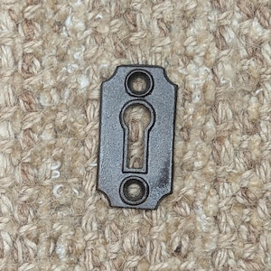 May include: A black metal keyhole cover with rounded edges and two small holes on either side of the keyhole.