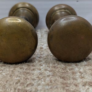 Könnte beinhalten: Zwei runde Türknöpfe aus Messing mit einer glatten, verwitterten Oberfläche. Jeder Knauf besteht aus einem kugelförmigen Griff, der an einer runden Grundplatte befestigt ist und Altersspuren sowie Patina auf der Metalloberfläche aufweist.