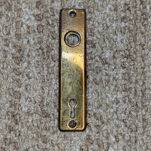 May include: A rectangular brass door escutcheon plate featuring a weathered, tarnished finish. The plate includes a circular opening at the top for a spindle, a keyhole opening at the bottom, and two small screw holes at the top and bottom ends.