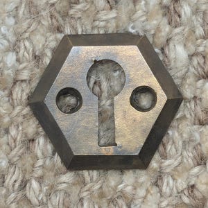 Op de afbeelding: Een zeshoekige messing sleutelgatbedekking met twee kleinere gaten aan weerszijden van het sleutelgat.