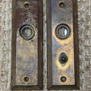 May include: Two antique brass door plates with a worn, aged finish. Each plate has a round hole and a smaller round knob. The plates are rectangular and have a dark brown wood frame.