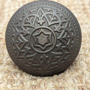 May include: A dark bronze-colored cabinet knob with an ornate, symmetrical design. The knob features a central six-pointed star surrounded by detailed floral and geometric patterns. The knob is round and has a slightly textured surface.