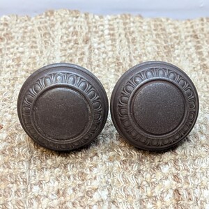 May include: Two dark brown metal cabinet knobs with a decorative, raised, circular design.