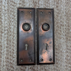 May include: Two rectangular door plates made of aged copper with a dark, weathered patina. Each plate features a central circular opening for a door knob, a lower keyhole, and four small screw holes located at the top and bottom corners.