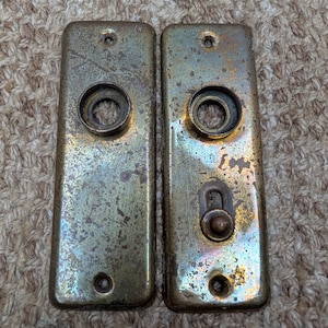 Peut inclure: Deux plaques de porte rectangulaires en laiton avec une finition patinée et ternie. Chaque plaque présente une ouverture circulaire centrale pour une poignée et deux petits trous de vis en haut et en bas. La plaque de droite inclut un loquet.