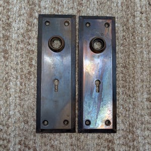 Könnte beinhalten: Zwei rechteckige Türschilder aus Metall mit einer dunklen, angelaufenen Oberfläche. Jedes Schild verfügt über eine kreisförmige Mittelöffnung für einen Türknauf, ein Schlüsselloch darunter und vier kleine Schraublöcher an den Ecken.