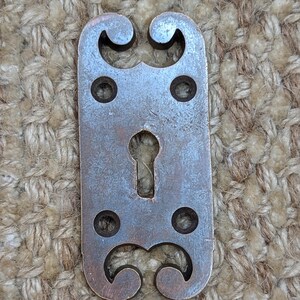 Pode incluir: Uma tampa de buraco de fechadura vintage em metal adornada, de forma retangular e com volutas decorativas. A tampa é de metal escuro envelhecido com uma pátina ligeiramente enferrujada.