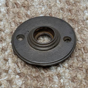May include: A dark gray, circular metal object with a central opening and two smaller holes. The object has a raised, tiered design around the central opening. The item appears to be a hardware component, possibly for a door or furniture.