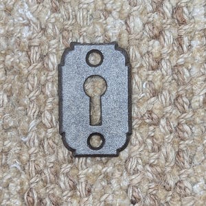 Könnte beinhalten: Ein silberner Schlüssellochausschnitt in einem dunkelbraunen Holzrahmen. Der Rahmen hat einen dekorativen Rand und zwei kleine Löcher oben und unten.