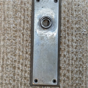 Pode incluir: Uma placa de porta de prata com um furo redondo no centro e quatro furos para parafusos nas bordas.