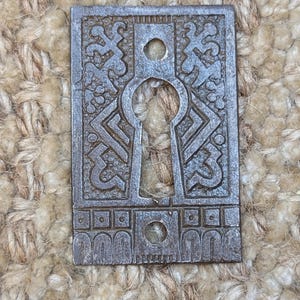 May include: A vintage keyhole cover made of metal with an intricate design. The cover is silver in color and has a keyhole in the center. The design features a repeating pattern of geometric shapes and swirls.