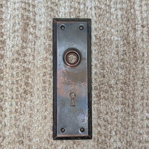 Op de afbeelding: Een rechthoekige metalen deurplaat met een verweerde bronsafwerking. De plaat is voorzien van een centrale ronde opening voor een deurklink, een sleutelgat eronder en vier kleine schroefgaten in de hoeken voor bevestigingsdoeleinden.