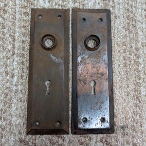 Könnte beinhalten: Zwei rechteckige Türschilder aus Metall mit einer verwitterten, dunkelbraunen Patina. Jedes Schild verfügt über eine kreisförmige Öffnung für den Türknauf oben, ein Schlüsselloch darunter und vier kleine Schraublöcher an den Ecken.