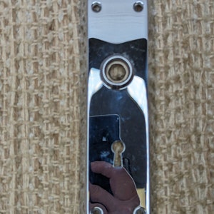 Może przedstawiać: Chromowana płytka drzwiowa z dziurką od klucza i dwoma zestawami otworów na śruby.