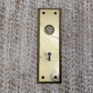 Könnte beinhalten: Eine rechteckige Türschildplatte aus Messing mit goldfarbenem Metallfinish, die eine zentrale kreisförmige Öffnung für einen Türknauf und ein unteres Schlüsselloch aufweist. Die Platte hat vier kleine Schraublöcher an den Ecken und Kratzer.