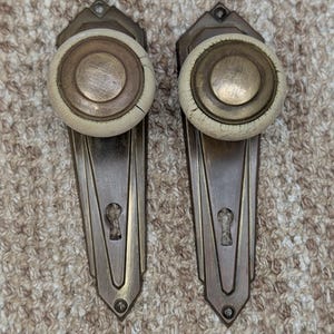 May include: A pair of door knobs with matching backplates, featuring a metallic bronze finish and circular off-white ceramic centers. The elongated backplates have a geometric shape with a central keyhole and visible screw holes at the top and bottom.