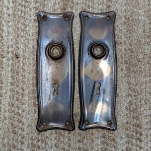 May include: Two rectangular metal door plates with a curved silhouette, featuring a central circular knob opening and a lower keyhole cutout. The plates have a weathered, tarnished finish with visible oxidation and small screw holes at each corner.