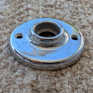 May include: A circular, polished chrome metal mounting plate featuring a raised, hollow center collar and two small, evenly spaced screw holes positioned on opposite sides of the central opening, designed for hardware installation.