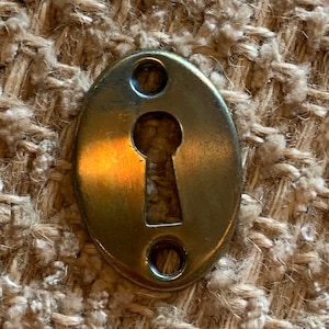 Könnte beinhalten: Ein ovaler Schlüsselschlitzabdeckung aus Messing. Die Abdeckung hat ein Schlüsselloch in der Mitte und zwei kleine Löcher an beiden Seiten.