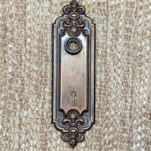Op de afbeelding: Een antieke bronzen deurplaat met een sleutelgat en een cirkelvormige opening voor een deurknop. De rechthoekige plaat heeft sierlijke bloemen- en krulwerkdetails. De deurplaat is waarschijnlijk gemaakt van gegoten metaal.