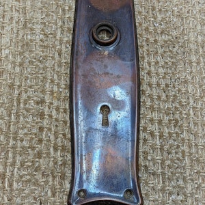 Puede incluir: Una placa de puerta rectangular vintage de cobre con una cerradura y un pomo redondo. La placa tiene una forma ligeramente curvada y una pátina desgastada y envejecida.